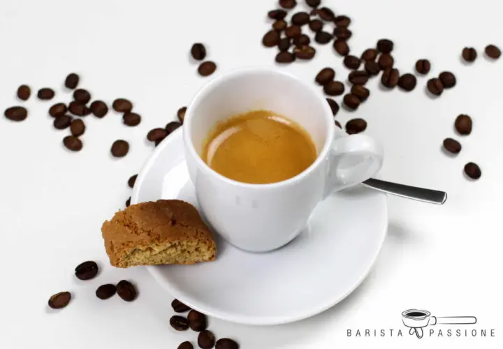 Ein weißer Becher mit Kaffee, daneben ein Cantuccini und Kaffeebohnen. Das ist ein Kaffee Lungo, ein längerer Espresso.