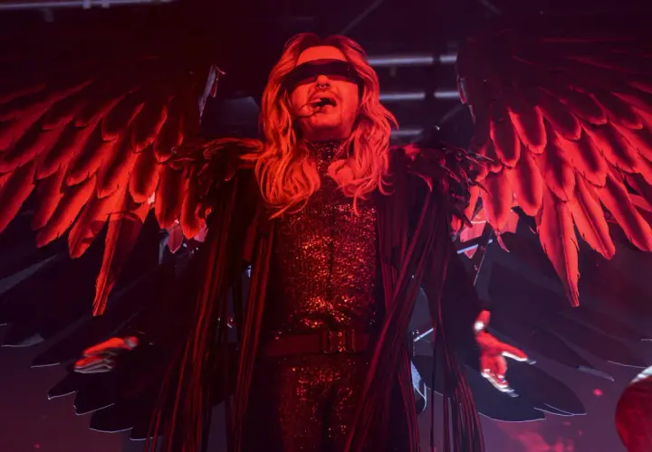 Bill Kaulitz z Tokio Hotel na koncercie w anielskich skrzydłach i błyszczącym stroju, w czerwonym świetle.