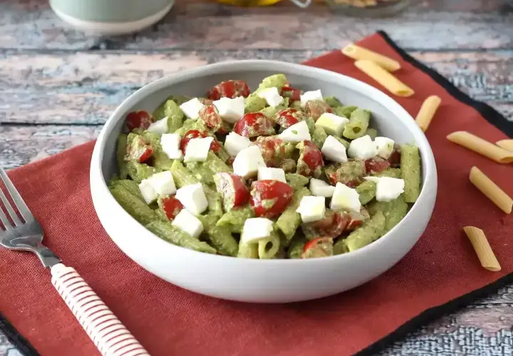 Pyszna pasta pesto z pomidorkami koktajlowymi i kostkami mozzarelli w białej misce, na czerwonej serwetce.
