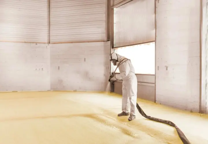 Un ouvrier en combinaison projette de la mousse polyuréthane sur le sol d'un entrepôt pour une isolation efficace.