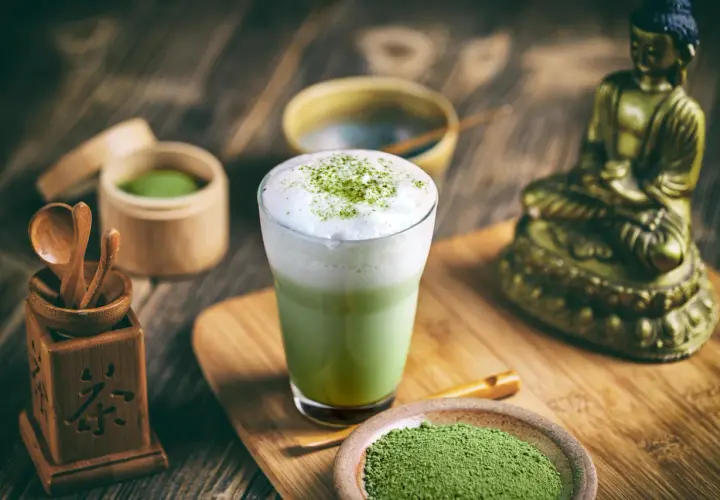 Szklanka zielonej herbaty matcha z pianką, obok naczynia z proszkiem matcha i bambusowe akcesoria.