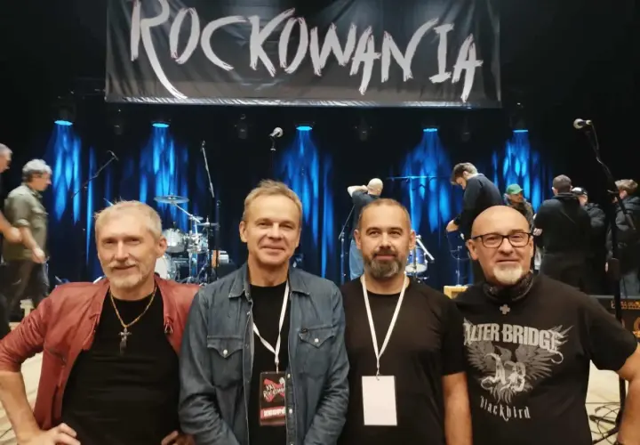 Czterech mężczyzn pozuje na tle sceny z banerem "ROCKOWANIA". Jeden z nich ma na sobie czerwoną kurtkę, inny dżinsową koszulę. W tle widać zespół i sprzęt. Idziemy do przodu dżem!