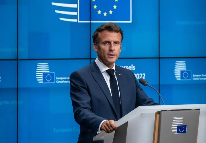Emmanuel Macron w garniturze przemawia za mównicą na tle niebieskiej ściany z logo Unii Europejskiej.