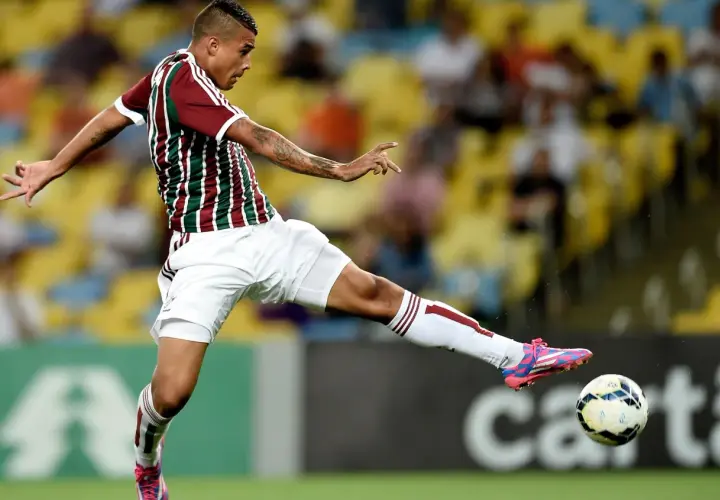 Piłkarz w koszulce Fluminense kopie piłkę na tle trybun.