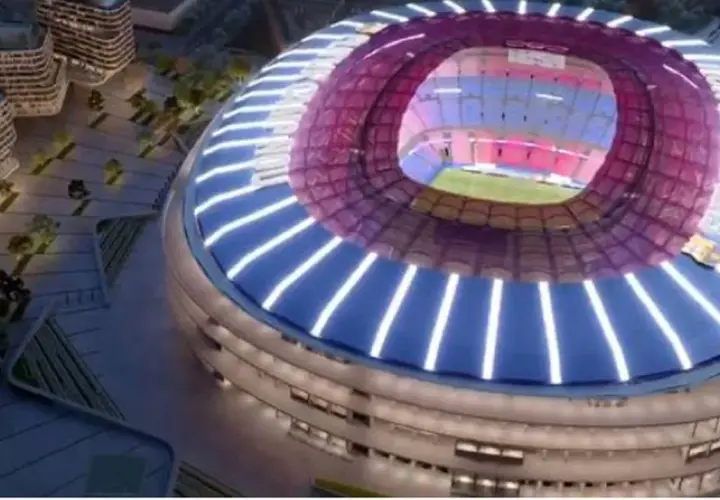 Nowe Camp Nou w nocnej odsłonie, z nowoczesną, oświetloną fasadą i dachem. Widać fragmenty otoczenia z budynkami i zielenią.