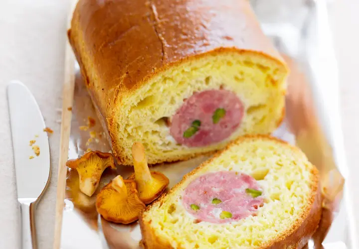 Un saucisson brioché doré, coupé en tranches, révèle sa garniture rosée. Des girolles et un couteau complètent cette gourmandise.