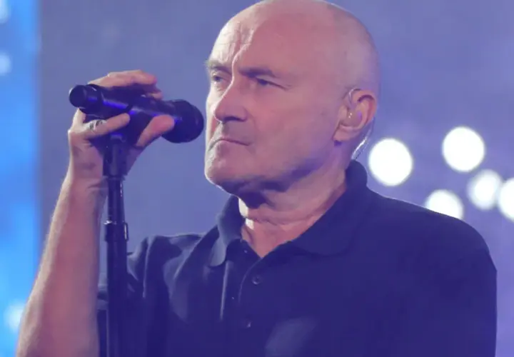 Phil Collins, wokalista zespołu Genesis, śpiewa do mikrofonu na tle niebieskiej sceny z białymi światłami.