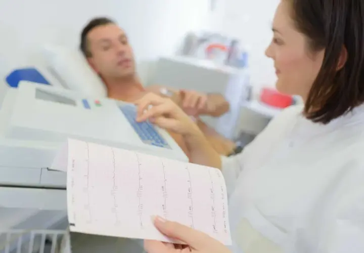 Pielęgniarka analizuje zapis EKG, który pokazuje, jakie są badania serca. Pacjent leży podłączony do aparatury.