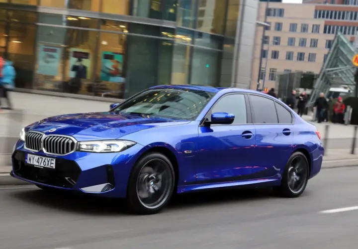 Błękitne BMW 330e mija nowoczesne budynki i wejście do metra.