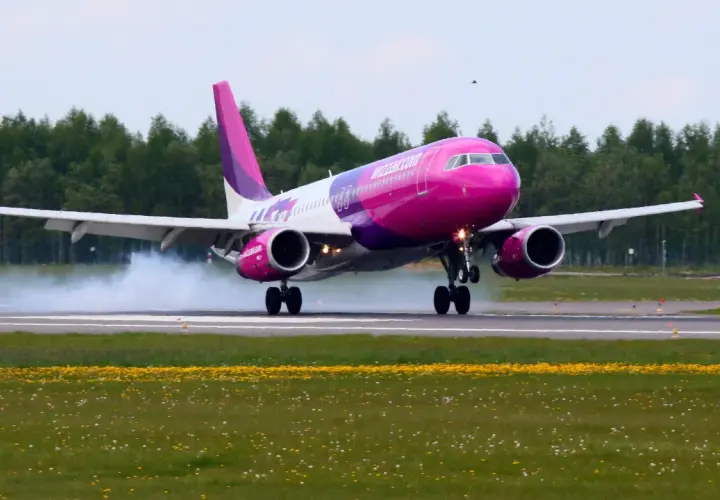 Różowy samolot Wizz Air ląduje na pasie startowym, unosząc dym.