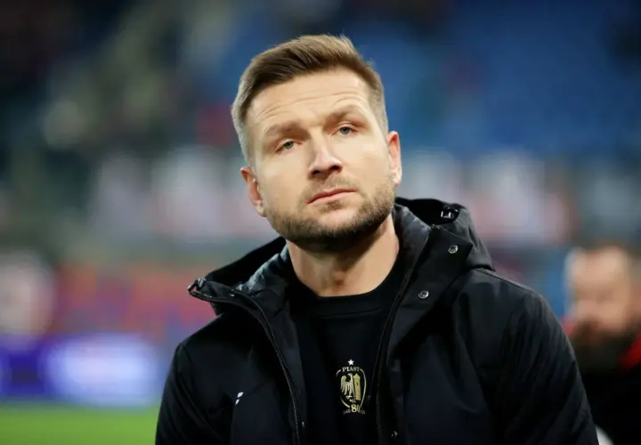 Mężczyzna w czarnej kurtce, z zarostem, w tle trybuny. To piast gliwice trener.