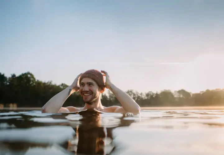Uśmiechnięty mężczyzna w czapce zanurzony w zimnej wodzie, czerpiący radość z metody wim hof oddychanie.