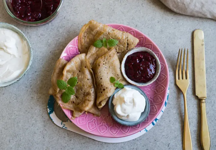 Glutenfria pannkakor med sylt och grädde på en rosa tallrik. En gaffel och kniv ligger bredvid.