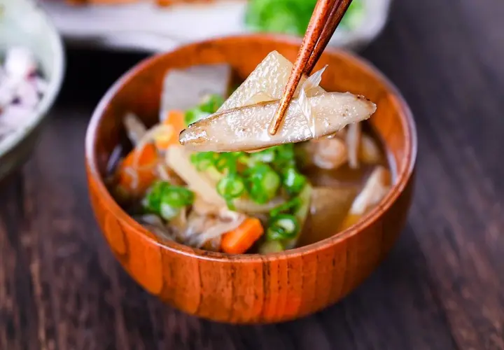 Chopsticks lift a piece of daikon from a steaming bowl of Japanese vegetable soup, filled with carrots, mushrooms, and green onions.
