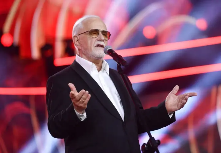 Jan Pietrzak w garniturze i okularach przeciwsłonecznych przemawia do mikrofonu na scenie.