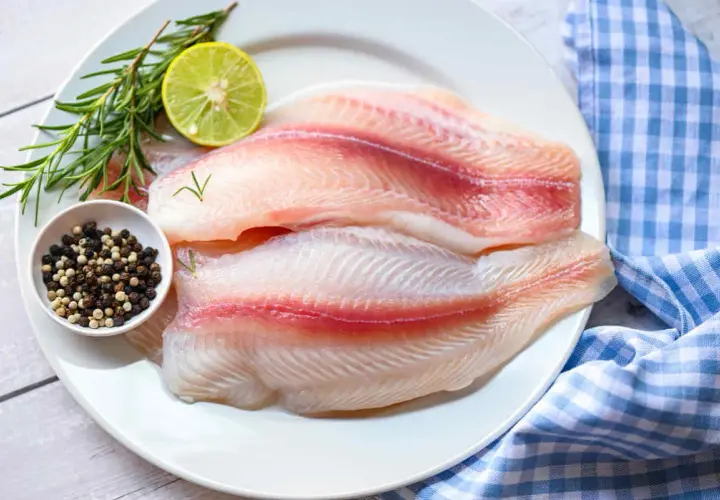 Dos filetes de tilapia frescos, listos para cocinar, acompañados de romero, limón y granos de pimienta.