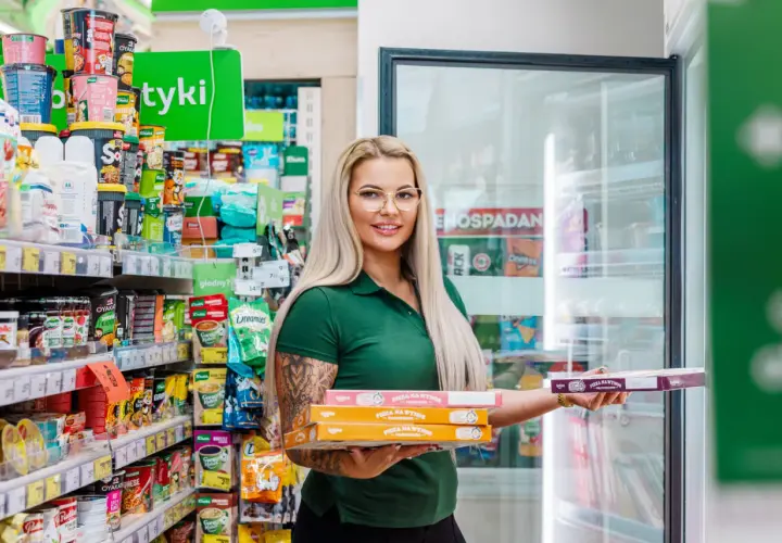 Kobieta w zielonej koszulce trzyma stos pudełek z pizzą na wynos w sklepie osiedlowym.