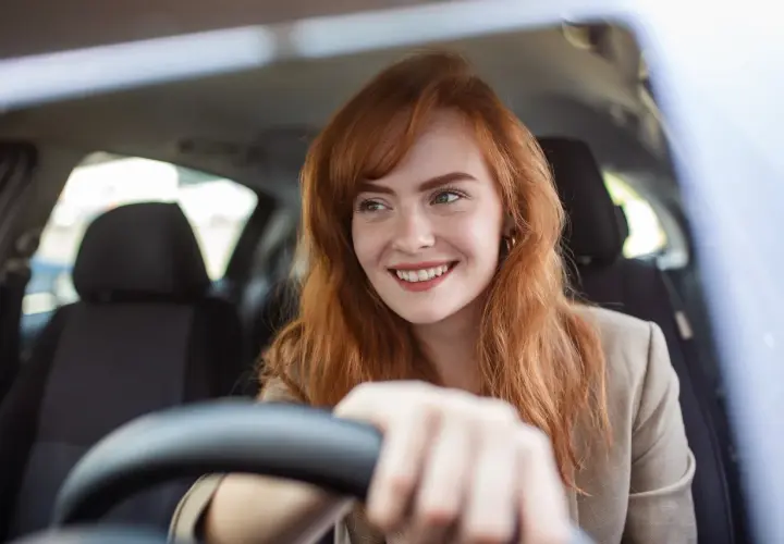 Donna sorridente al volante, con capelli rossi e lentiggini. Pensa alla sua assicurazione giornaliera auto per un viaggio sereno.