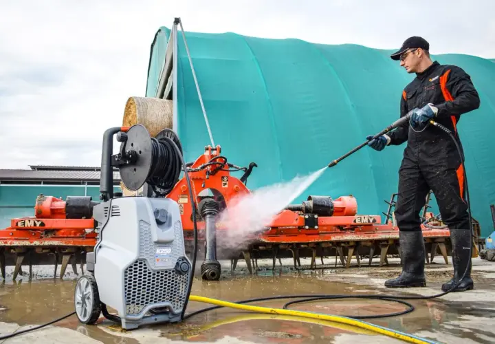 Hombre con traje de trabajo y botas limpiando maquinaria agrícola con hidrolimpiadora. Aprende como usar hidrolimpiadora para dejar todo impecable.