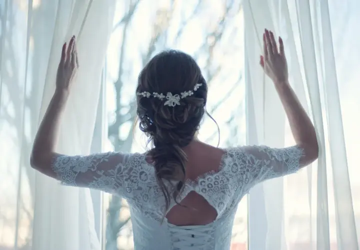 Elegancka fryzura dla świadkowej na wesele, z delikatną ozdobą we włosach, w białej koronkowej sukni, odsłaniająca zasłony.