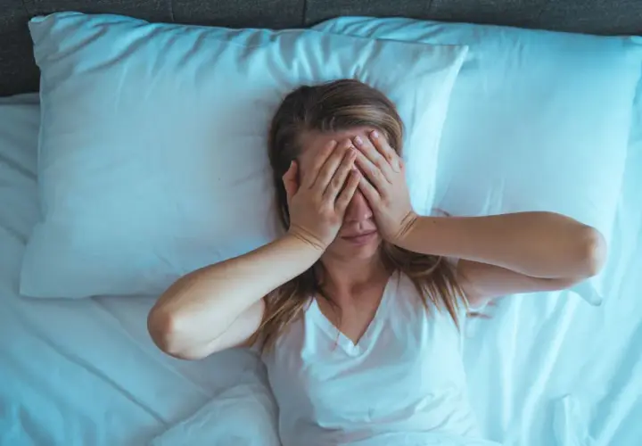 Mujer en la cama, con las manos cubriendo sus ojos. Son las 4 de la mañana y no puedo dormir, la luz azulada ilumina la escena.