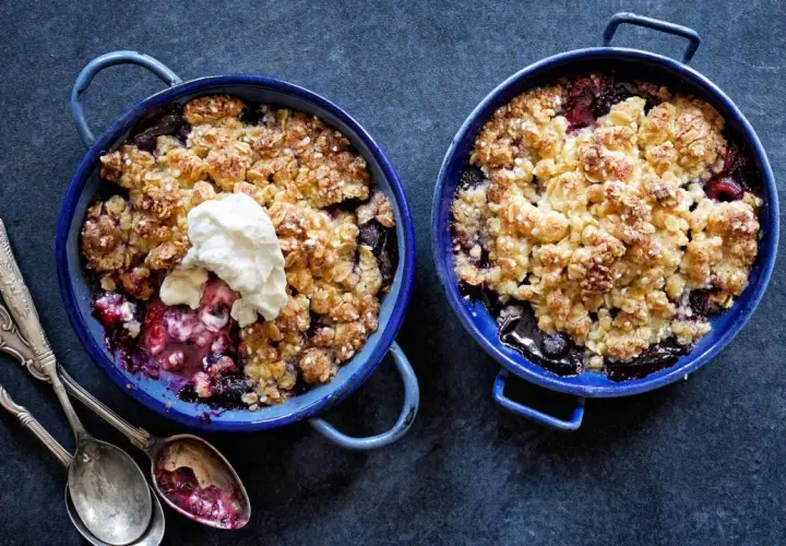 Två blåa ugnsformar med glutenfri smulpaj fylld med bär och toppad med grädde. Serveras med antika skedar.