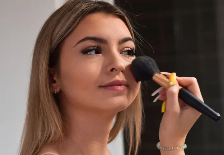 Młoda dziewczyna z długimi blond włosami nakłada puder pędzlem. Jej makijaż jest subtelny, podkreślający naturalne piękno, w stylu "clean girl".