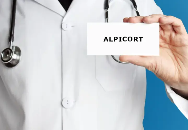Lekarz w białym kitlu prezentuje kartkę z napisem ALPICORT. Stetoskop wisi na jego szyi.