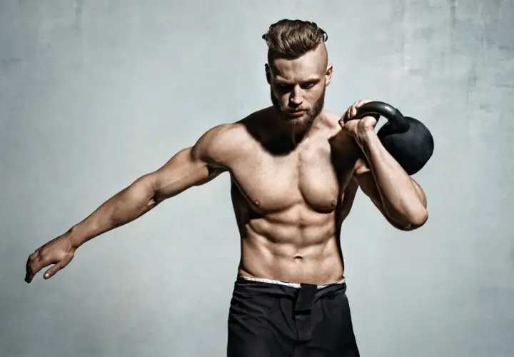 Mężczyzna z wyrzeźbionym brzuchem i kettlebell na ramieniu. Pokazuje, jak wejść na masę, trenując siłowo.