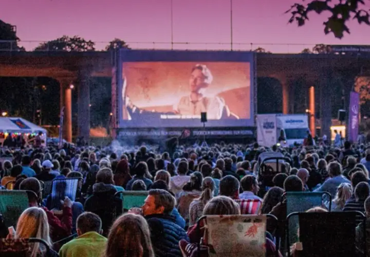 Stor publik njuter av sommarbio Stockholm under en lila kvällshimmel. En film visas på en stor duk framför tält och träd.