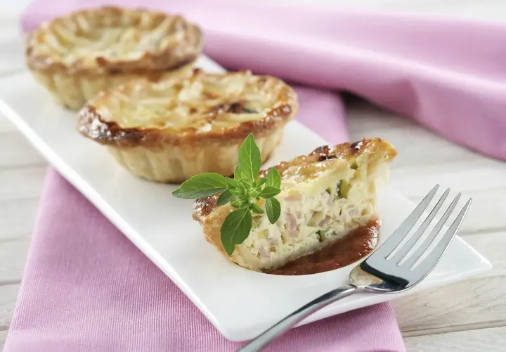 Trois mini quiches sans pâte garnies d'une tranche coupée, sur une assiette blanche avec une feuille de basilic.