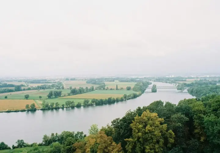 Szeroka rzeka na tle pól i lasów, z mostem w oddali.