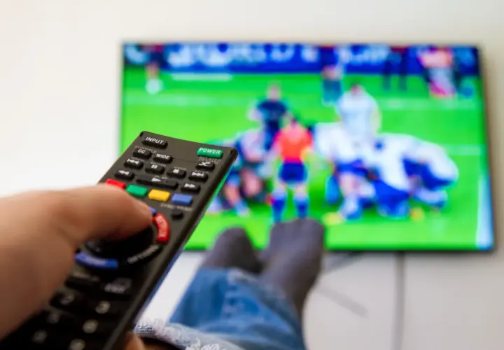 Dłoń trzymająca pilot do telewizora, na ekranie mecz piłki nożnej.