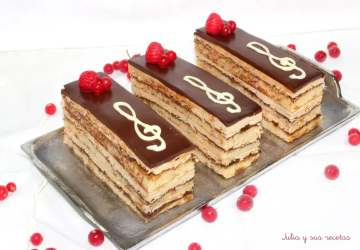 Tres porciones de pastel ópera, con capas de bizcocho y crema, cubiertas de chocolate y decoradas con clave de sol y frutos rojos.