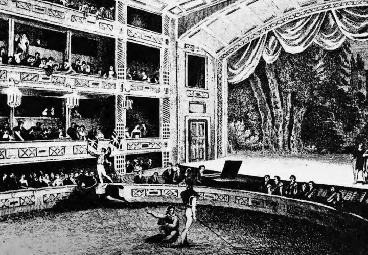 Widownia wypełnia wielopoziomowe loże, a na arenie trwa spektakl. To scena z życia teatru średniowiecznego, pełna widzów i aktorów.