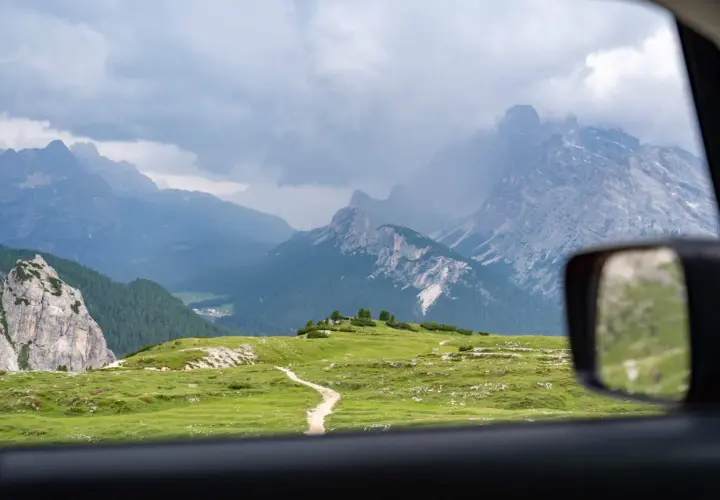 Vue depuis une voiture en Italie, traversant des prairies verdoyantes et des montagnes majestueuses sous un ciel nuageux.