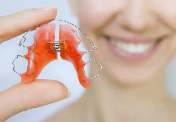 Dłoń trzyma pomarańczowy, wieloczynnościowy aparat dentystyczny. W tle uśmiechnięta twarz.