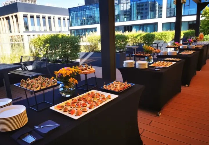 Elegancki catering Warszawa na imprezę plenerową. Stoły z przekąskami i napojami na tarasie z widokiem na nowoczesne budynki.