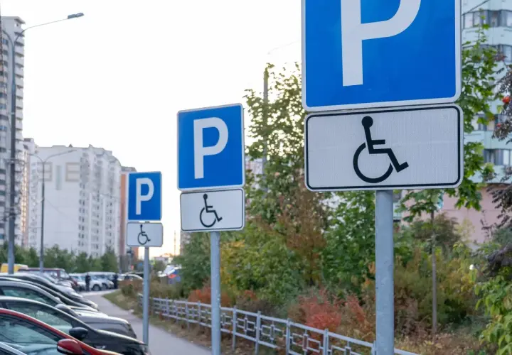 Znaki informują, komu przysługuje karta parkingowa. Na parkingu stoją samochody, a w tle widać budynki mieszkalne.