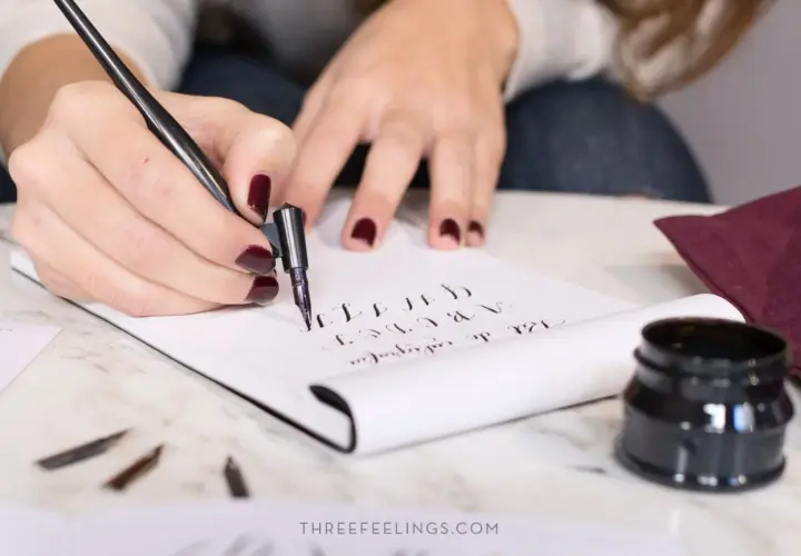 Manos practicando lettering con pluma y tinta, creando letras cursivas en un cuaderno.