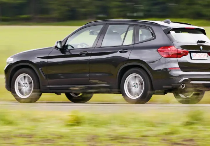 Ciemnoszary BMW G01 pędzi po wiejskiej drodze, rozmazując zielone pola i drzewa w tle.