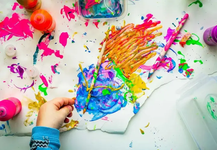 Kinderhand malt mit bunten Farben auf Papier, umgeben von Farbtöpfen und Pinseln.