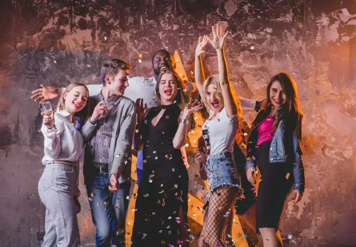 Grupa przyjaciół świętuje w klubie, sypiąc konfetti. Inspiracja, jak się ubrać do klubu: sukienka, jeansy, topy, marynarka.