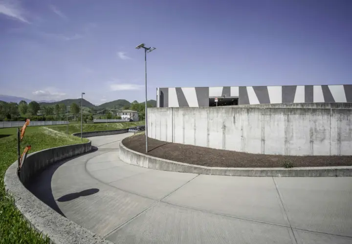 Ampia rampa in cemento fai da te, curva dolcemente verso un edificio moderno con facciata a strisce bianche e grigie, circondata da verde e colline.