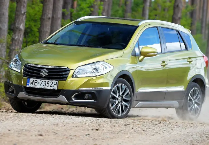 Zielony Suzuki S-Cross na drodze leśnej, wzbudzający pozytywne suzuki s-cross opinie. Kurz spod kół świadczy o jego terenowych możliwościach.
