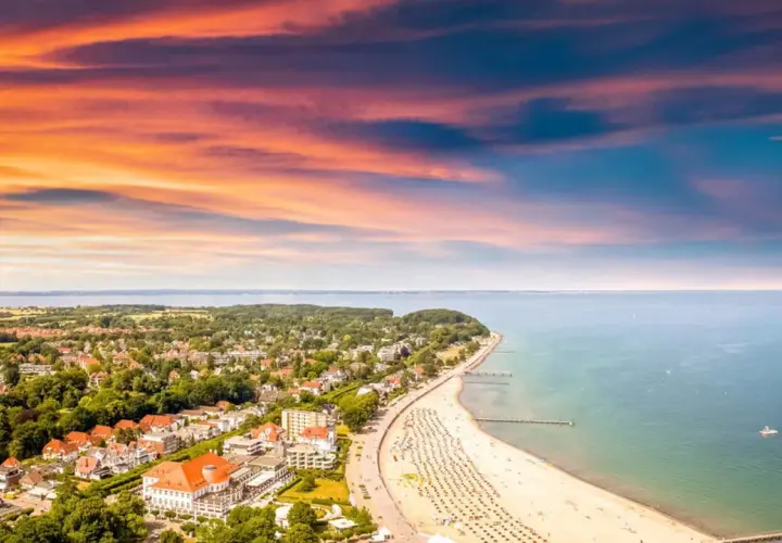 Ein perfekter tagesausflug ostsee: Sonnenuntergang über der Ostsee, belebter Strand mit Liegen und ein Küstenort mit roten Dächern.