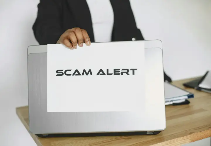 Ostrzeżenie przed oszustwami na Vinted. Ktoś trzyma kartkę z napisem "SCAM ALERT" nad laptopem.
