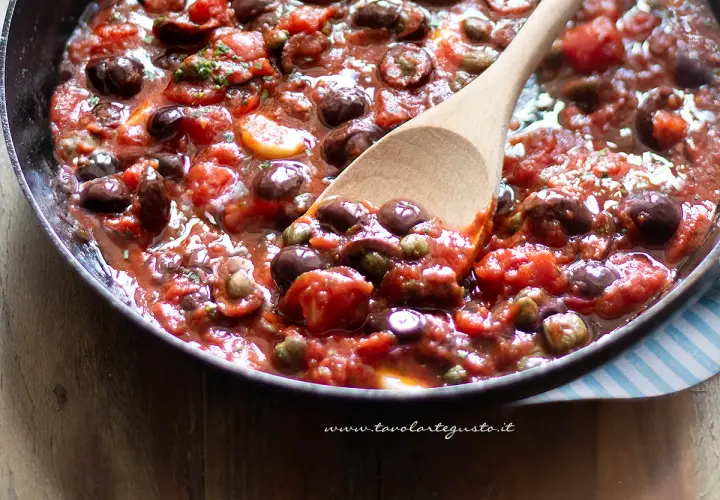 Un cucchiaio di legno mescola un sugo ricco e saporito, pieno di pomodori, olive nere e capperi. È la base perfetta per una pasta alla puttanesca.
