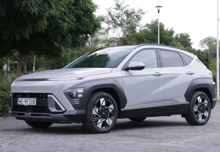 Nowy Hyundai Kona Hybrid w kolorze jasnoszarym, zaparkowany na brukowanym placu, otoczony zielenią.