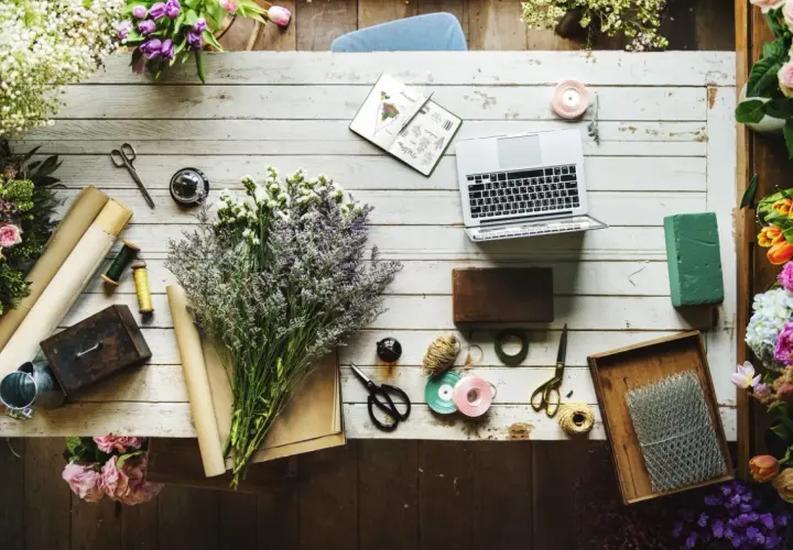 Kwiaty, laptop i akcesoria florystyczne, jak nożyczki, wstążki i sznurek, tworzą inspirujący warsztat. Widać też materiały do pakowania, przypominające **faszynę**.